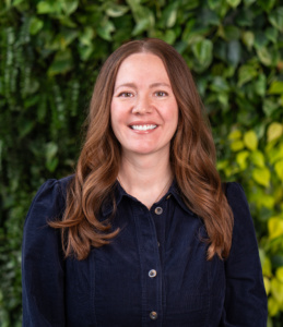 Woman with long wavy auburn hair wearing a dark blue buttoned dress standing against a leafy green background.