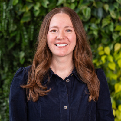 Woman with long wavy auburn hair wearing a dark blue buttoned dress standing against a leafy green background.