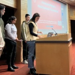 Six students present in front of a group with a slide presentation in the background