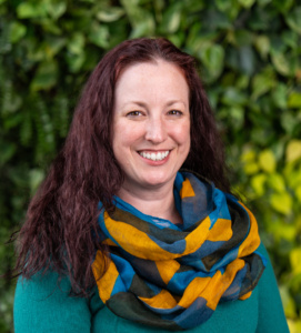 Woman with long curly brown hair wearing a teal sweater and a colorful blue, yellow, and black patterned scarf against a leafy green background.