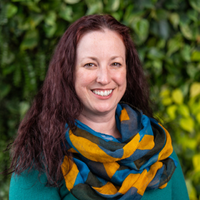 Woman with long curly brown hair wearing a teal sweater and a colorful blue, yellow, and black patterned scarf against a leafy green background.