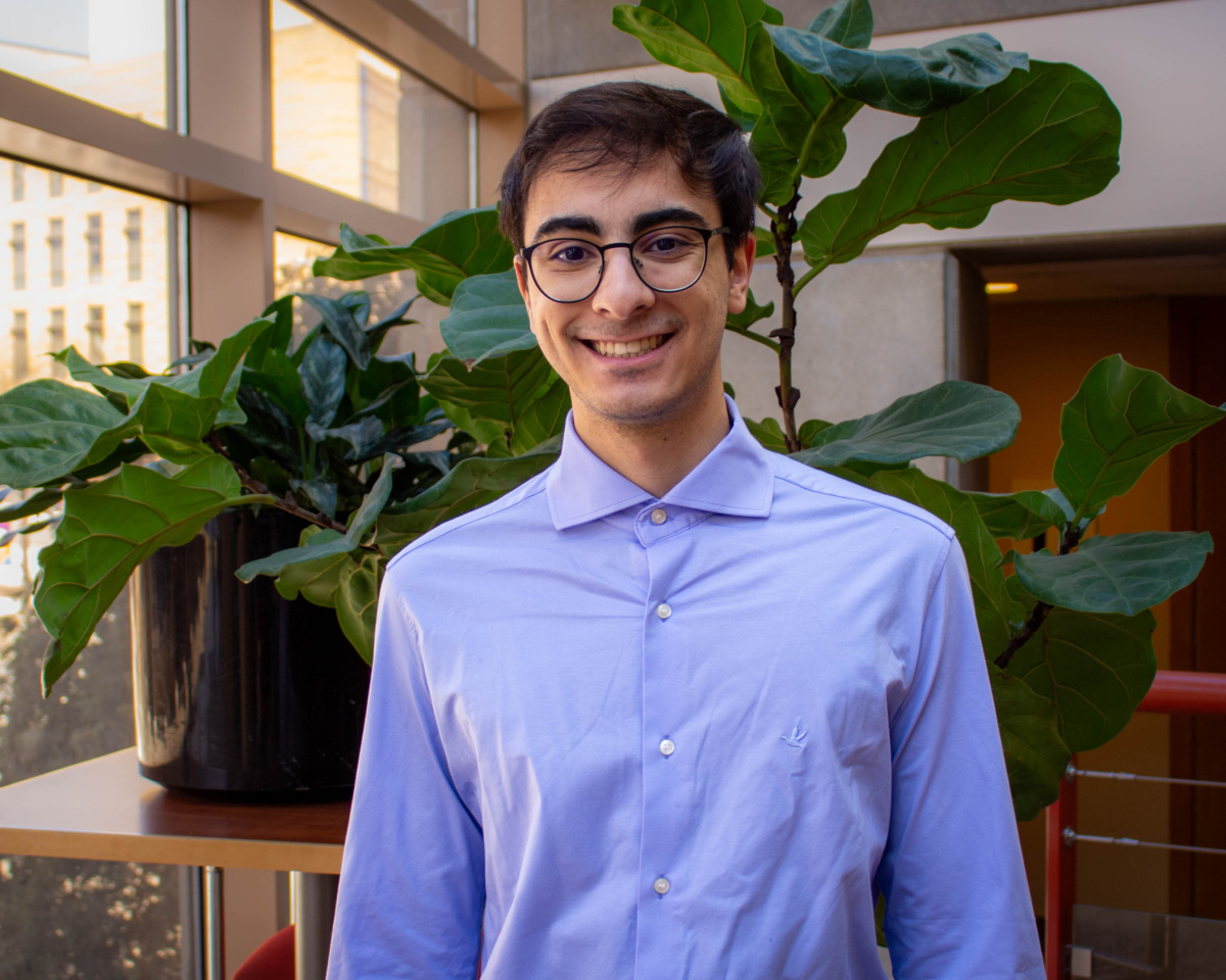 A man with brown hair wearing a blue shirt, Lucas Sernik, stands in front of plants