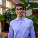 A man with brown hair wearing a blue shirt, Lucas Sernik, stands in front of plants