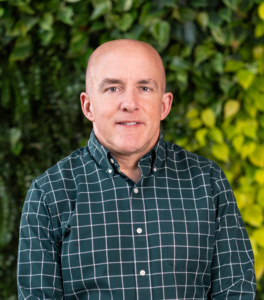 Man wearing a dark green checkered button-up shirt standing with leafy green foliage in the background.