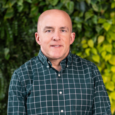 Man wearing a dark green checkered button-up shirt standing with leafy green foliage in the background.