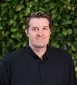 Man wearing a black button-up shirt standing in front of a green leafy background.