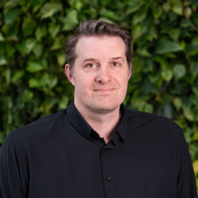 Man wearing a black button-up shirt standing in front of a green leafy background.