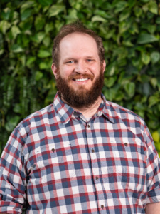 Man wearing a red, white, and blue plaid button-up shirt standing outdoors with green leafy plants in the background.