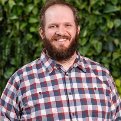 Man wearing a red, white, and blue plaid button-up shirt standing outdoors with green leafy plants in the background.