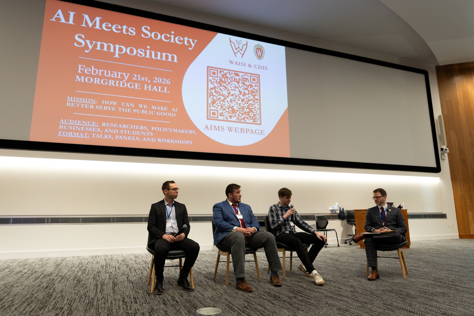 Speakers sit at the front of the Hello, World! auditorium in Morgridge Hall during the AI Meets Society Symposium.