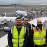 Three airport workers wearing high-visibility vests standing on a platform with Alaska Airlines planes and airport equipment in the background.