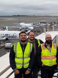 Three airport workers wearing high-visibility vests standing on a platform with Alaska Airlines planes and airport equipment in the background.