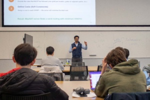 Tannu explaining quantum concepts on a whiteboard to attentive students in a classroom setting.