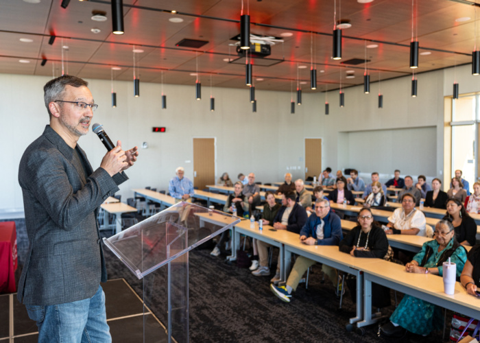 Remzi Arpaci-Dusseau addresses event attendees sitting at long tables in a modern room.