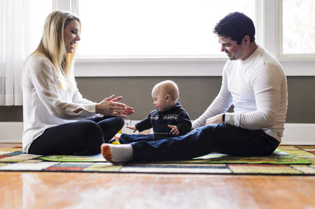 Parents sitting on a colorful rug playing and interacting with their baby in a bright room with large windows.