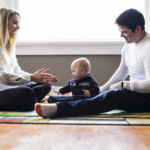 Parents sitting on a colorful rug playing and interacting with their baby in a bright room with large windows.